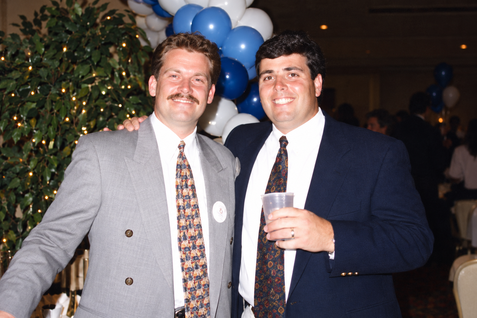 Two men stand side by side beneath the reunion balloons. Color interpretation.
